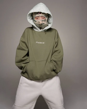 Model wearing an olive green hoodie, light grey quilted hood, camo balaclava, and white sweatpants in a studio.