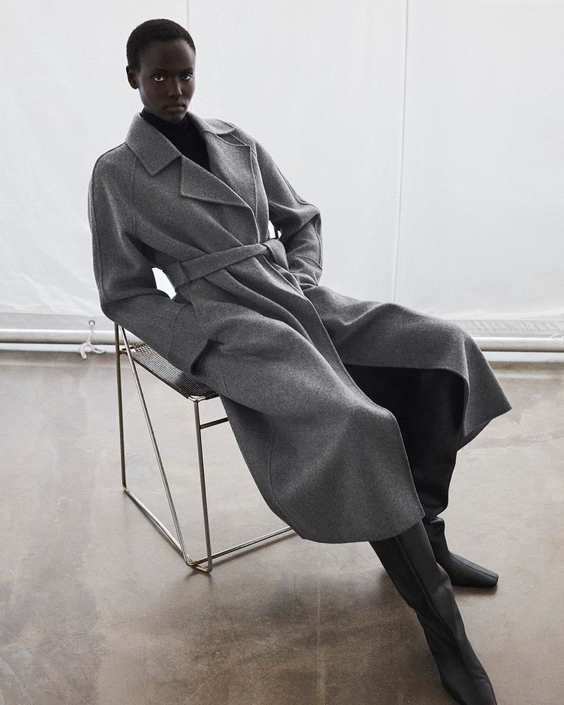 An African American woman sits on a wire chair in a grey belted coat and black turtle neck sweater with knee-high boots in a studio.