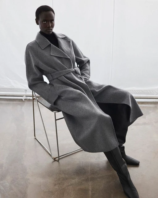 An African American woman sits on a wire chair in a grey belted coat and black turtle neck sweater with knee-high boots in a studio.