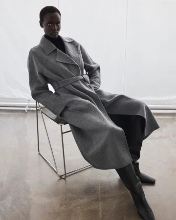 An African American woman sits on a wire chair in a grey belted coat and black turtle neck sweater with knee-high boots in a studio.