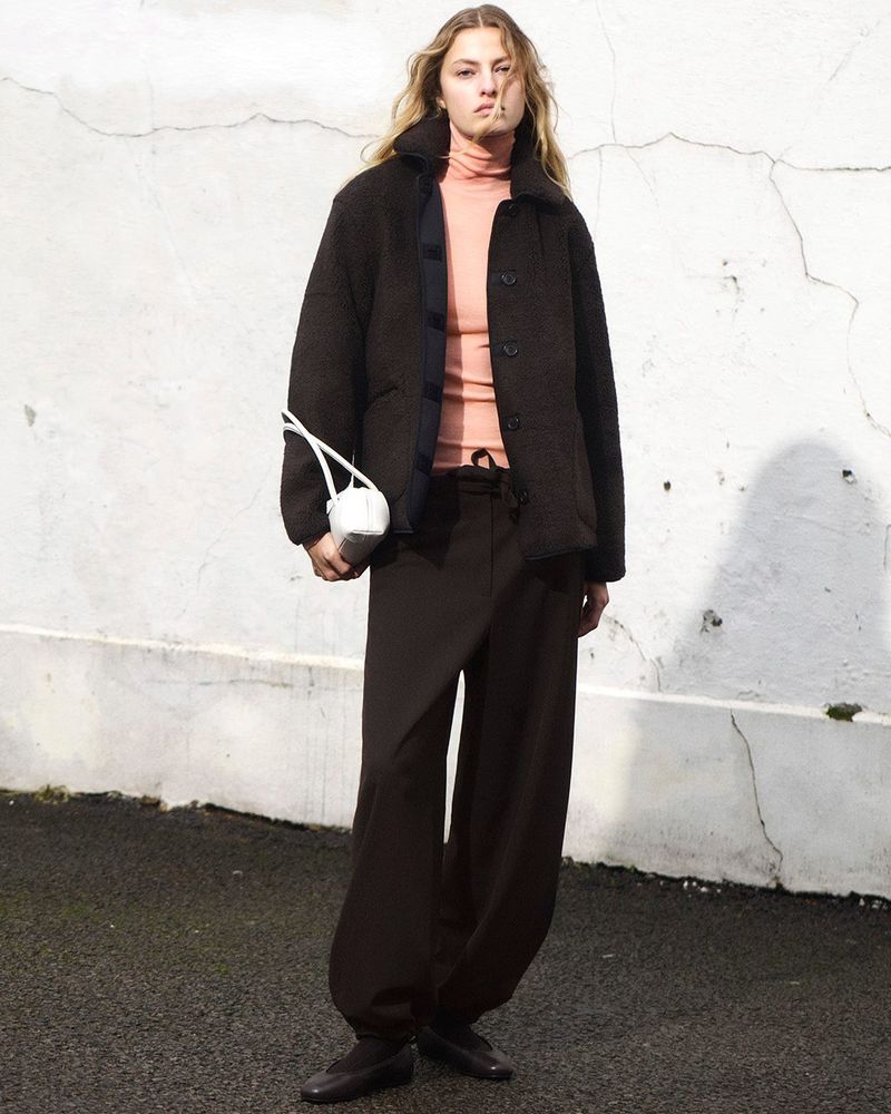 A female fashion model poses in a brown jacket and pants against a textured white wall, holding a white bag with white handles.