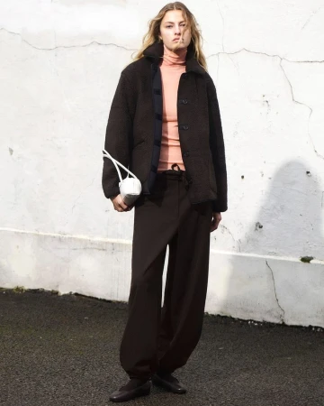 A female fashion model poses in a brown jacket and pants against a textured white wall, holding a white bag with white handles.