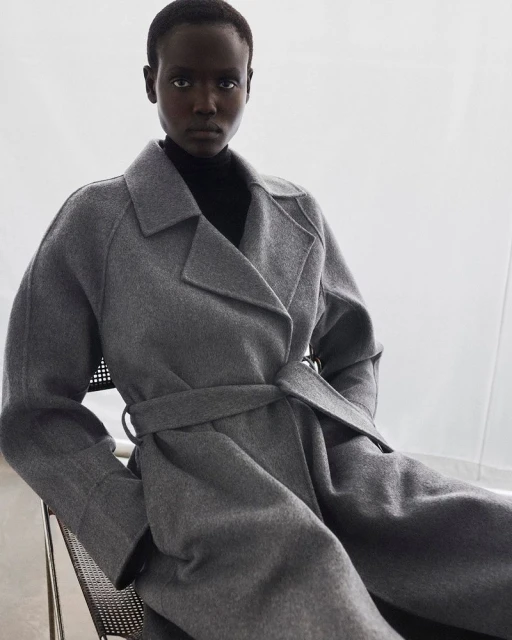 A solitary African-American woman in a grey belted coat seated in a light studio, capturing a blend of fashion and contemplative mood.