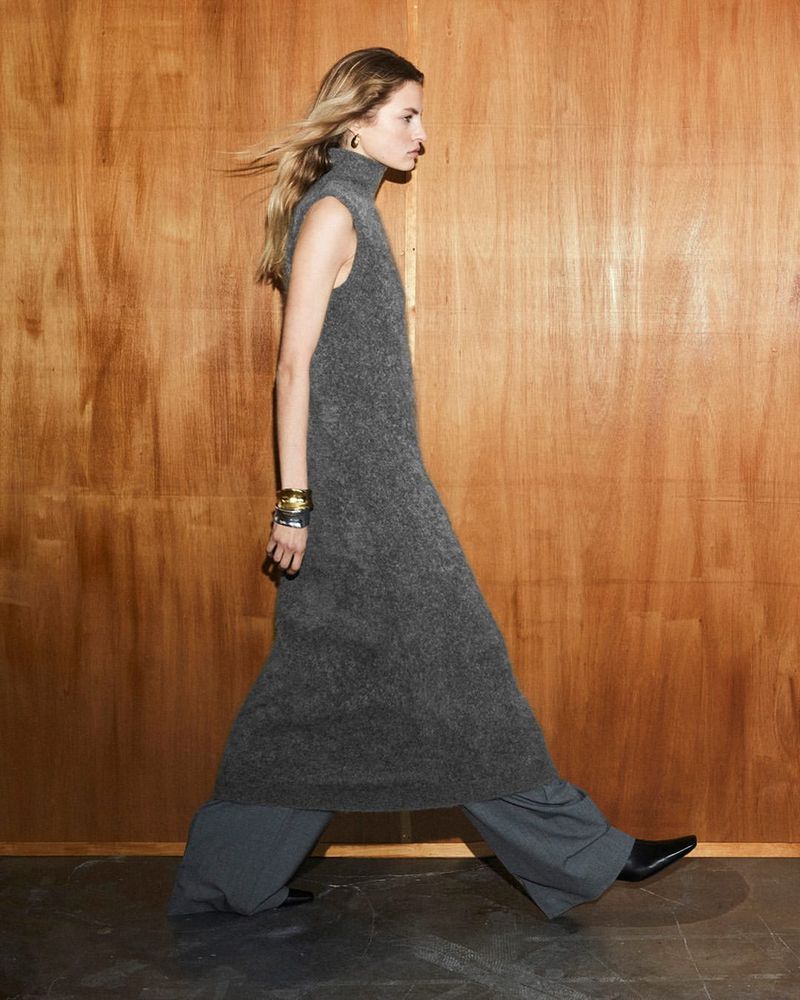 A tall blonde adult female in a gray knit dress walks against a wood panel backdrop, accessorized with gold earrings and silver bangles.