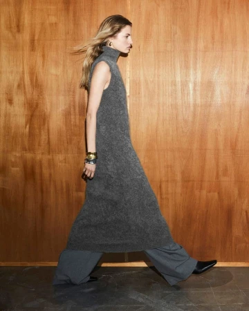 A tall blonde adult female in a gray knit dress walks against a wood panel backdrop, accessorized with gold earrings and silver bangles.