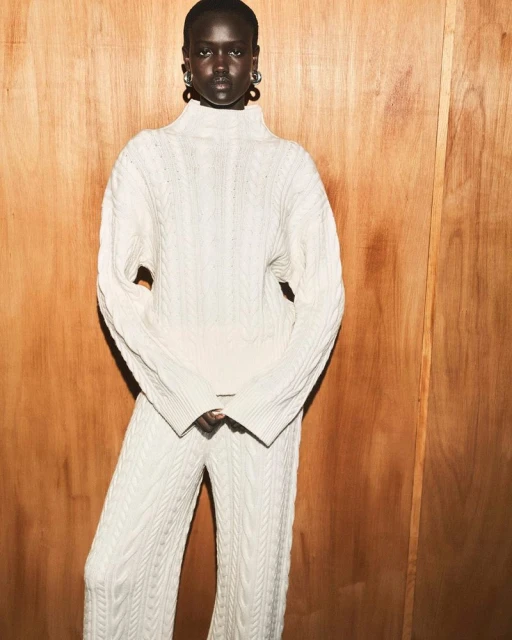 Woman poses in a cream-colored cable knit sweater and matching pants in front of a wood paneled wall. Fashion editorial shot.