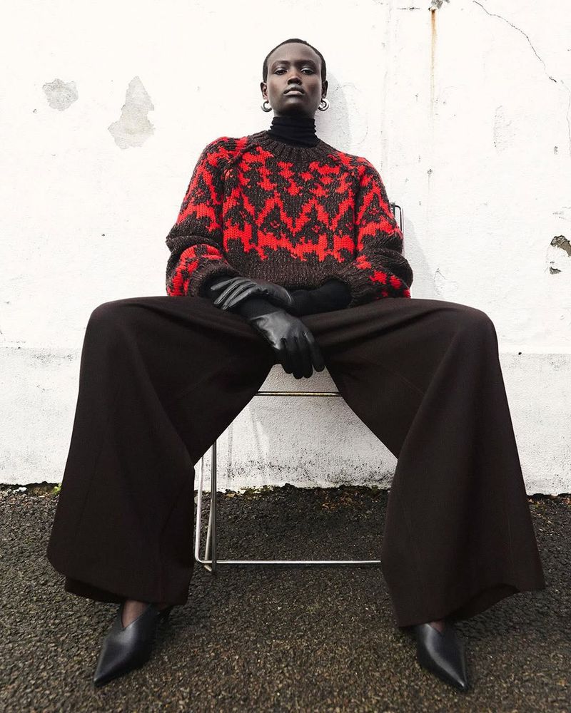 A stylish portrait of an African woman in an outdoor fashion shoot, wearing a patterned sweater, wide-legged pants, and black heels, sitting on a chair.