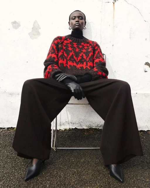 A stylish portrait of an African woman in an outdoor fashion shoot, wearing a patterned sweater, wide-legged pants, and black heels, sitting on a chair.