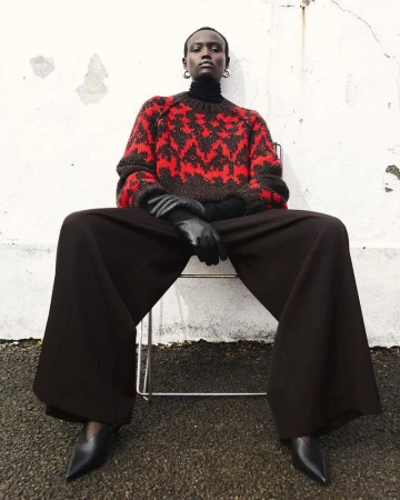 A stylish portrait of an African woman in an outdoor fashion shoot, wearing a patterned sweater, wide-legged pants, and black heels, sitting on a chair.