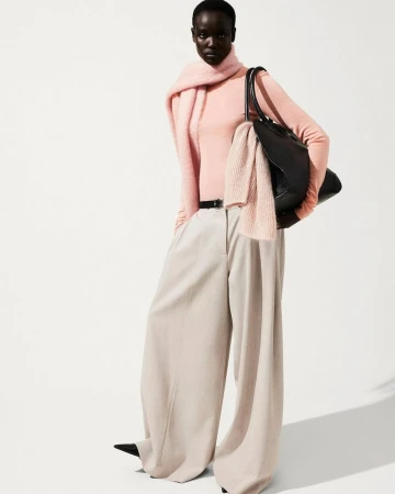 Model in pink sweater and wide-leg trousers holding black leather bag against a white seamless background