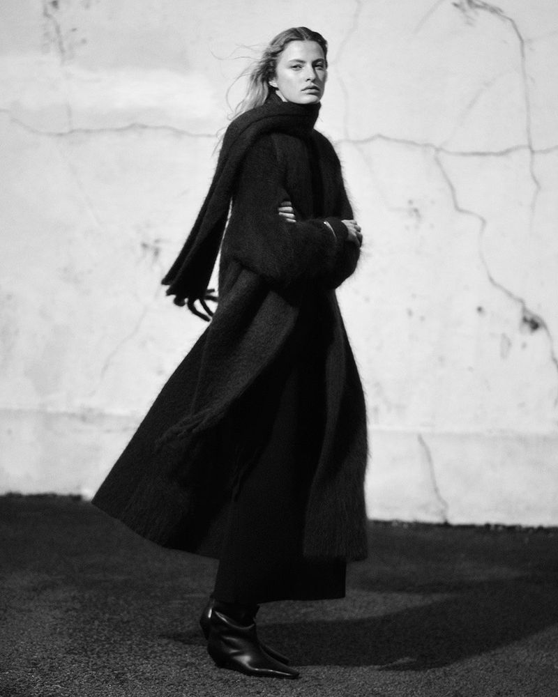 A woman in a long, dark coat and scarf poses outdoors against a textured wall, creating a monochrome fashion portrait.