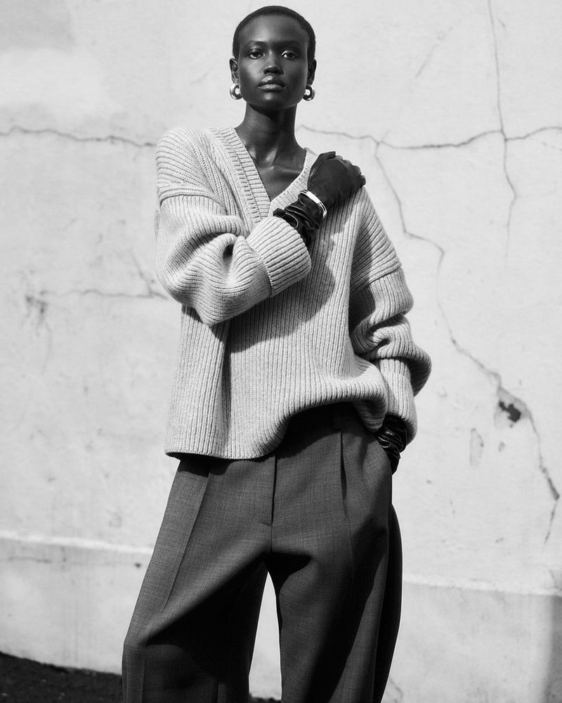 Monochrome portrait of a Black woman wearing a neutral sweater, gray slacks, earrings, and black gloves posing against a textured wall.