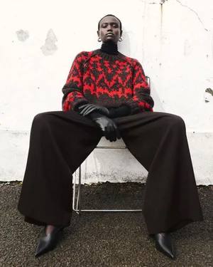 Model wearing a patterned red and black sweater and wide dark trousers sitting on a chair against a white wall.