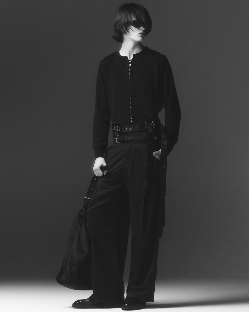 Monochrome fashion shot of a person with long hair wearing a dark cardigan, wide-leg trousers with straps, and carrying a black bag.