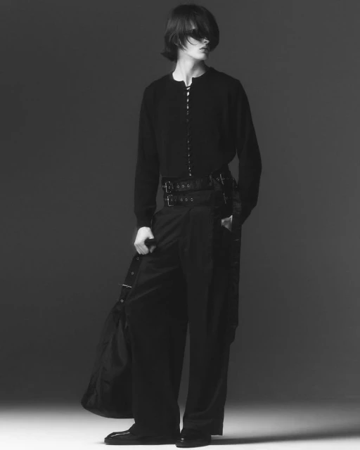 Monochrome fashion shot of a person with long hair wearing a dark cardigan, wide-leg trousers with straps, and carrying a black bag.