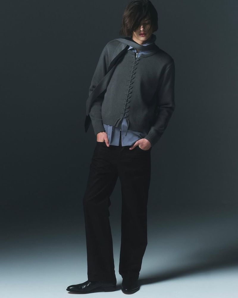 Fashionable young adult poses in a studio wearing a gray knitted cardigan, blue button-down shirt, black pants, and black dress shoes.