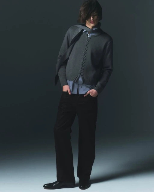 Fashionable young adult poses in a studio wearing a gray knitted cardigan, blue button-down shirt, black pants, and black dress shoes.
