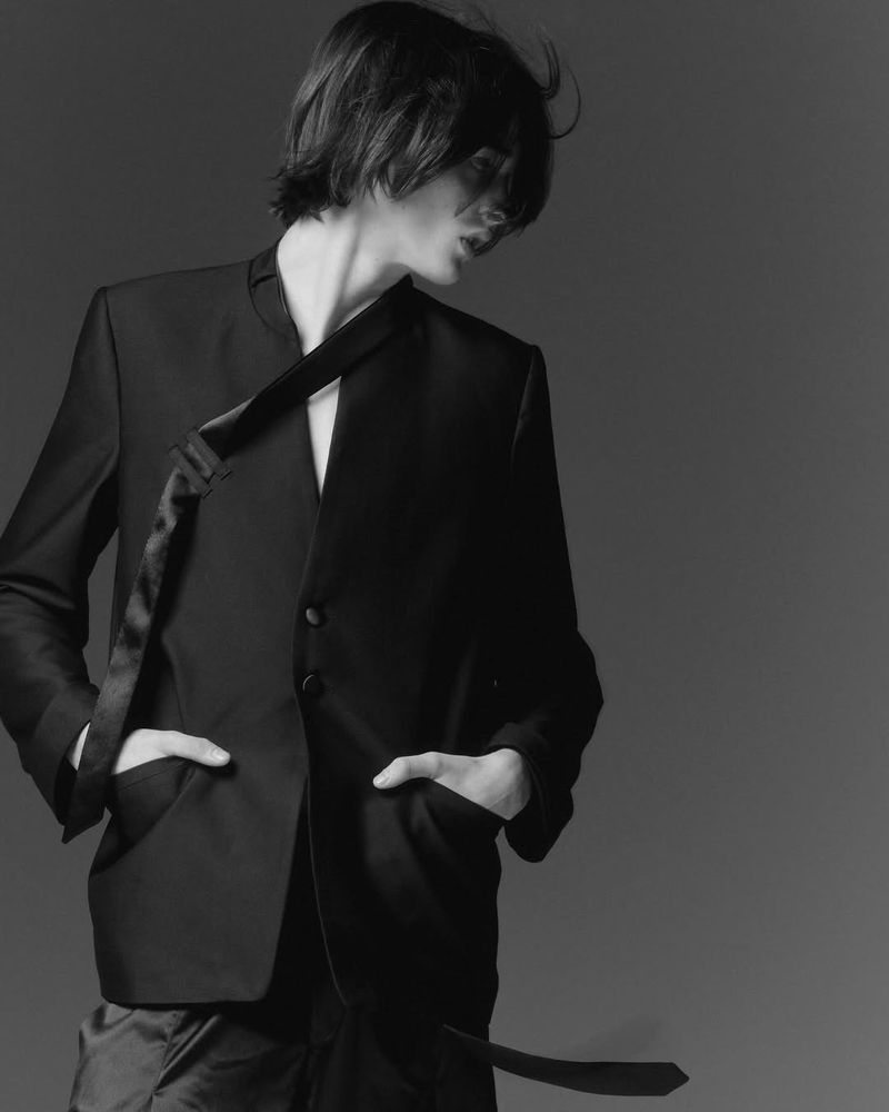 Model wearing a black jacket with hands in pockets against a grey studio backdrop