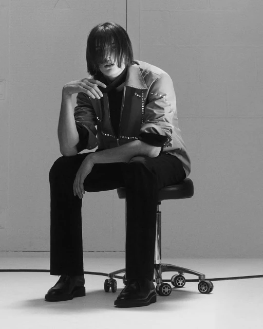 A person seated on a rolling stool, clad in a studded jacket, black top and trousers, and black boots, poses against a grey backdrop.