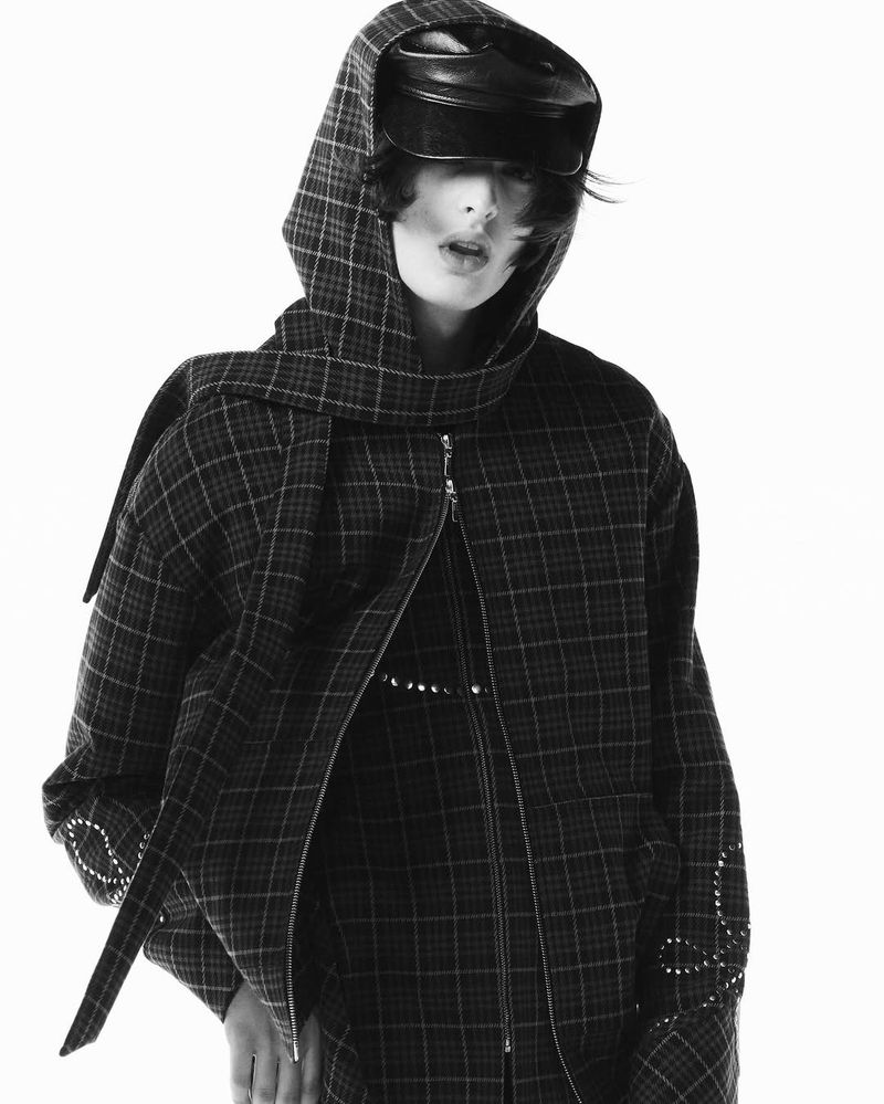 Monochrome portrait of a person wearing a plaid hooded jacket with a shiny black hat against a white background in a fashion editorial style.