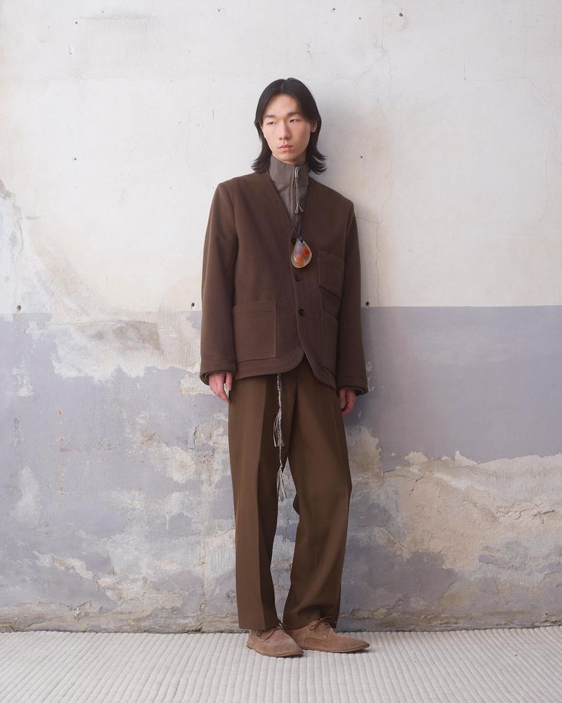 Adult male in a brown blazer and pants stands against a textured wall. Full shot of a person in earth-toned clothing in a neutral setting.