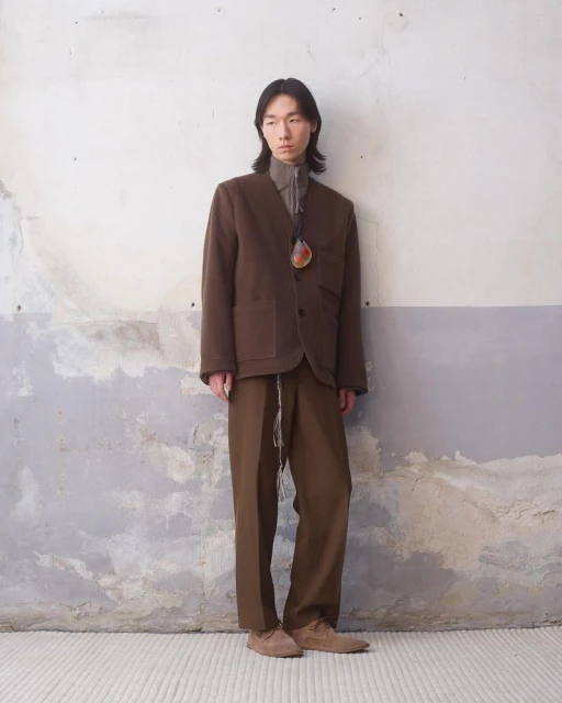 Adult male in a brown blazer and pants stands against a textured wall. Full shot of a person in earth-toned clothing in a neutral setting.