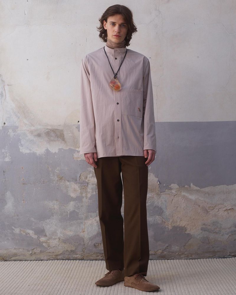 A slender man in a neutral-striped shirt, brown trousers, and suede shoes stands against a weathered wall, showcasing fashion apparel.