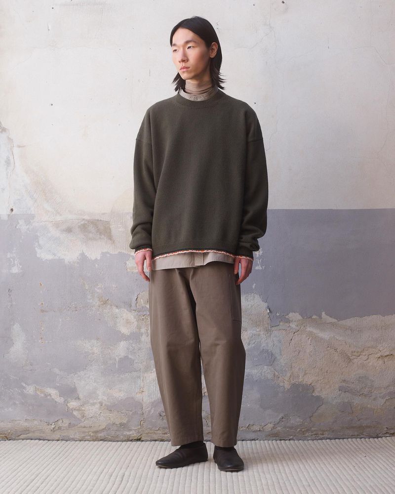 An Asian male model stands against a textured wall, wearing an olive green sweatshirt, khaki pants, and black shoes in a studio shot.