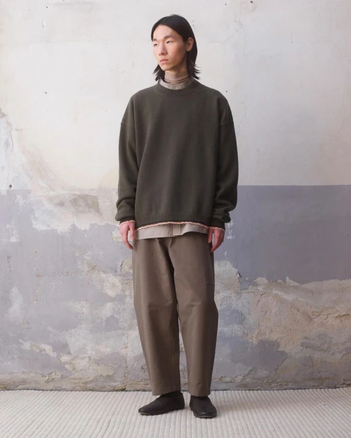 An Asian male model stands against a textured wall, wearing an olive green sweatshirt, khaki pants, and black shoes in a studio shot.