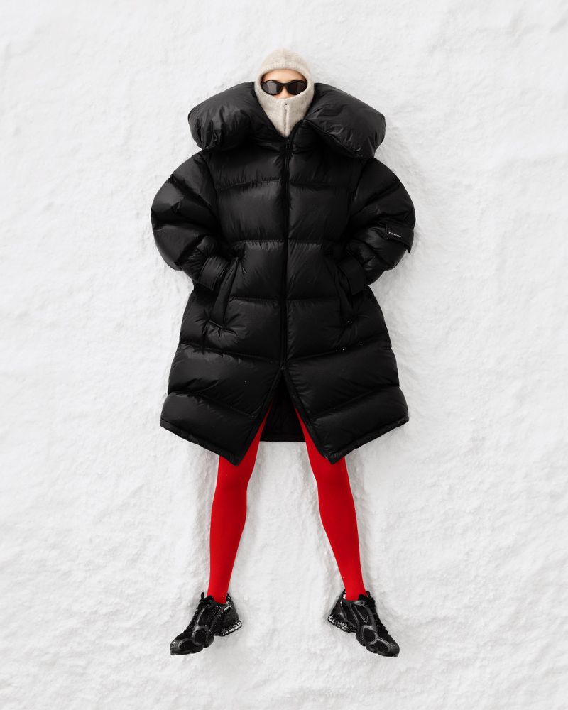 A person wearing a black puffer coat, sunglasses, and bright red tights laying on a white surface that looks like snow.