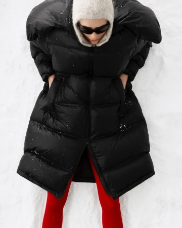 Overhead shot of a woman lying in snow, wearing a black puffer coat, bright red tights, sunglasses, and a knit balaclava.
