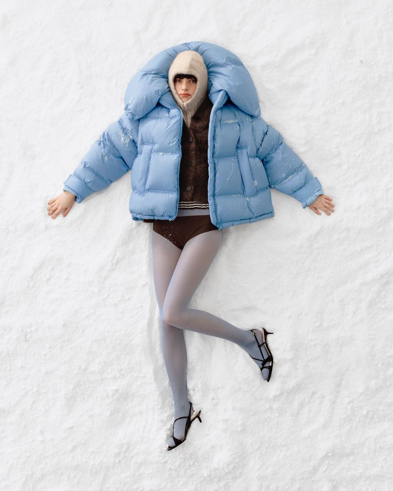 Fashion model in blue puffer jacket and balaclava lies on white textured surface, styled with tights and heels, eye-level, studio lighting.