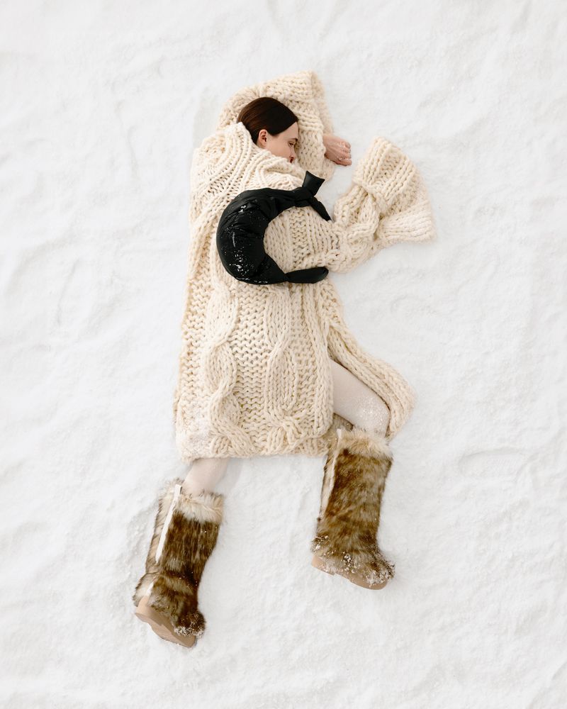 Overhead shot of a woman lying on a white textured surface, covered by a long cream cable-knit cardigan and wearing furry boots.