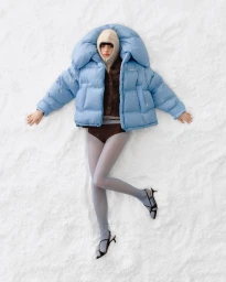 Model lying in the snow wearing a blue puffer jacket, balaclava, tights, and slingback heels