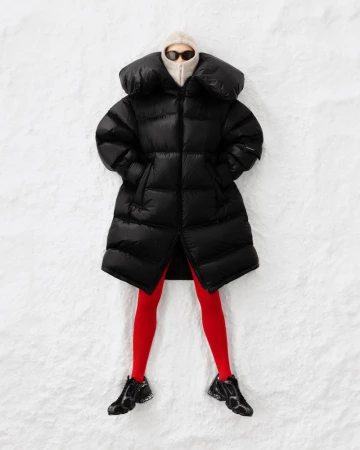 Model wearing a black oversized puffer coat, red tights, and dark sunglasses lying on a snow surface