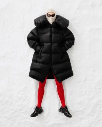 Model wearing a black oversized puffer coat, red tights, and dark sunglasses lying on a snow surface