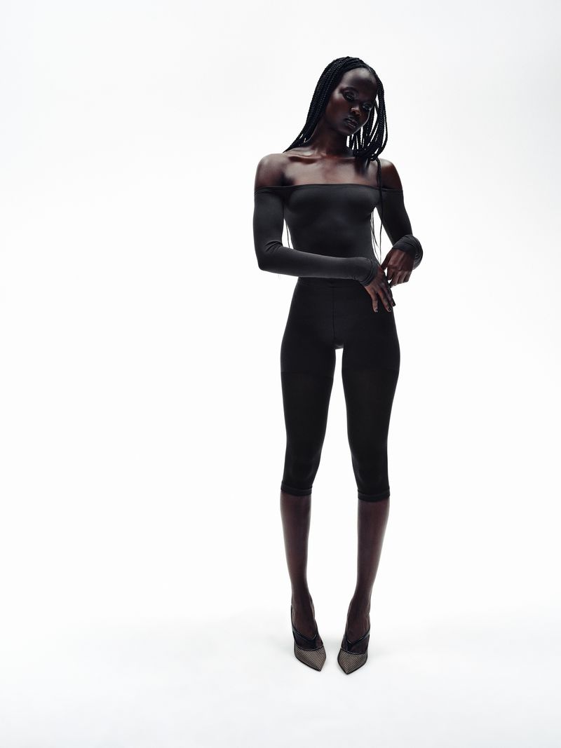 Elegant Black female fashion model poses in form-fitting dark top and Capri leggings, wearing pointed heels on a white background.