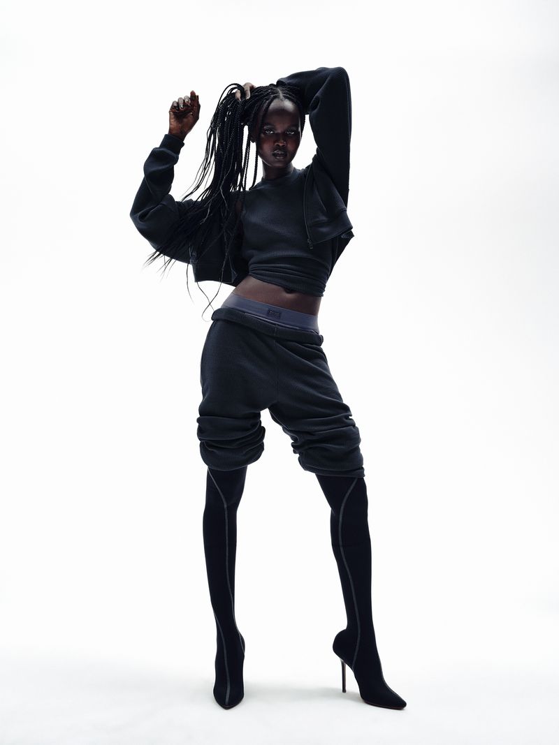 Dark-skinned woman in a dark athletic outfit strikes a pose in a studio with a bright white backdrop, creating a high-contrast fashion image.