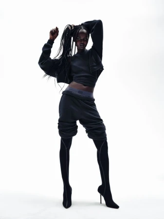 Dark-skinned woman in a dark athletic outfit strikes a pose in a studio with a bright white backdrop, creating a high-contrast fashion image.