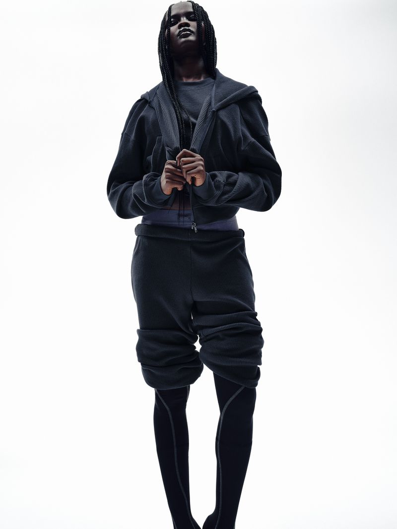 Woman in dark athleisure wear posing in a bright, minimalist studio, showcasing unique textile textures and modern fashion elements.