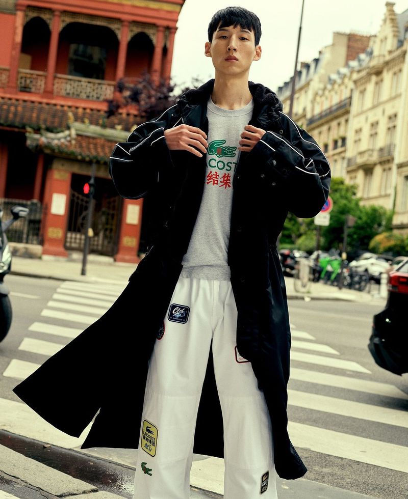 A young adult man poses in an urban setting while wearing a long black coat, grey shirt, and patterned white pants near city buildings.