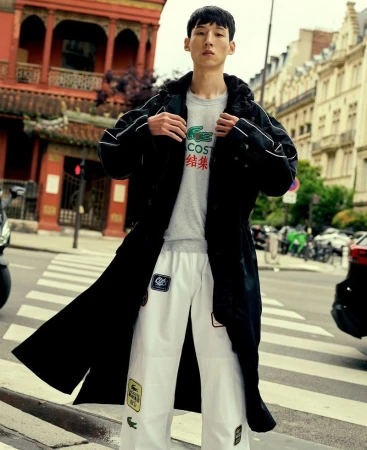 A young adult man poses in an urban setting while wearing a long black coat, grey shirt, and patterned white pants near city buildings.