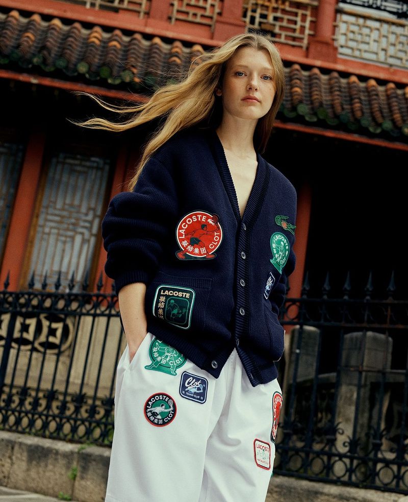 Fashion shot featuring a blonde woman in a Lacoste cardigan and white pants standing in front of a traditional building.