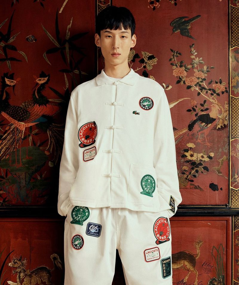 Asian male fashion model in Lacoste-themed apparel with logo patches against a red oriental floral backdrop in an indoor setting.