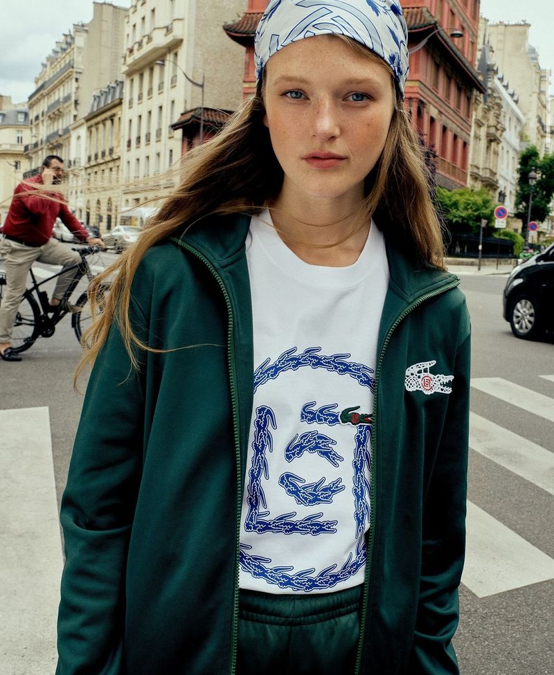 A young adult woman in a teal Lacoste tracksuit and headscarf stands on a city street with buildings in the background.
