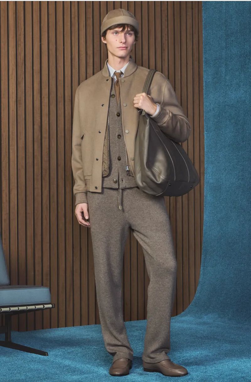 Model standing in a tonal beige wool jacket and trousers holding a large dark leather tote bag against a wood panel and blue backdrop.