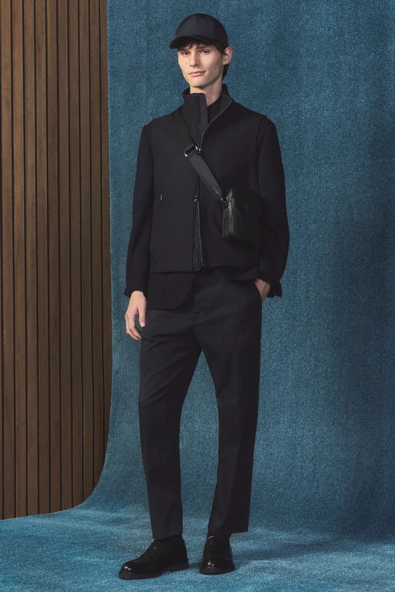 A full shot of a slender male model in a black cap, black vest, dark pants and dress shoes, standing in front of a textured backdrop.