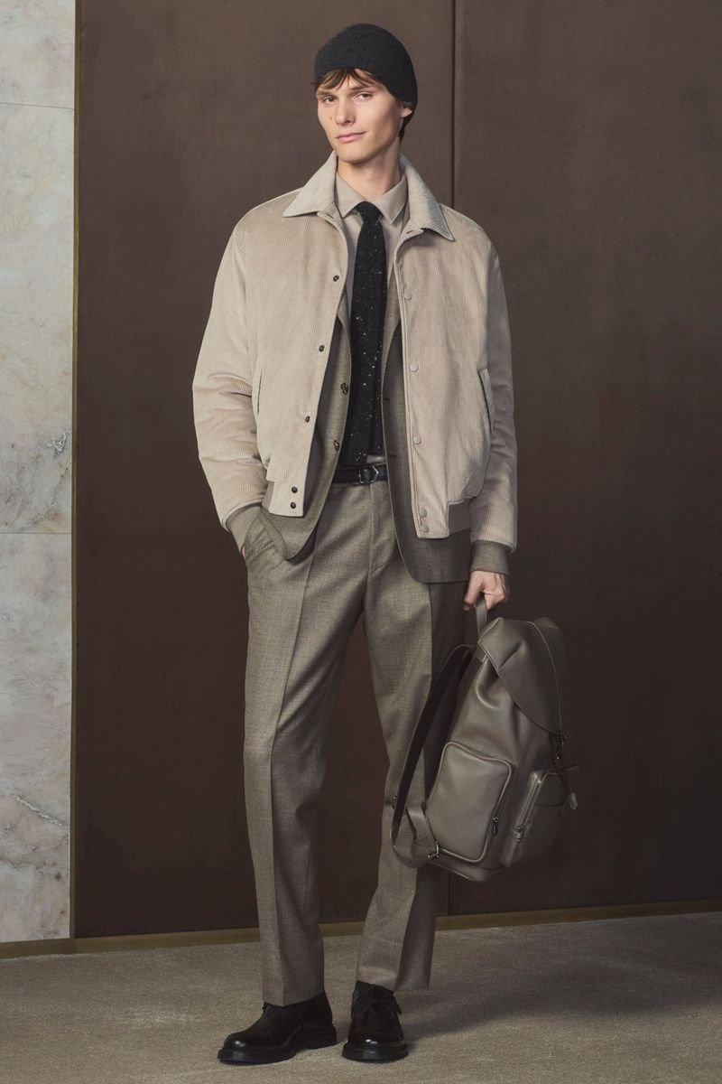 Full-length studio shot of a male model wearing a sand-colored jacket, grey plaid suit, black beanie, and holding a backpack.