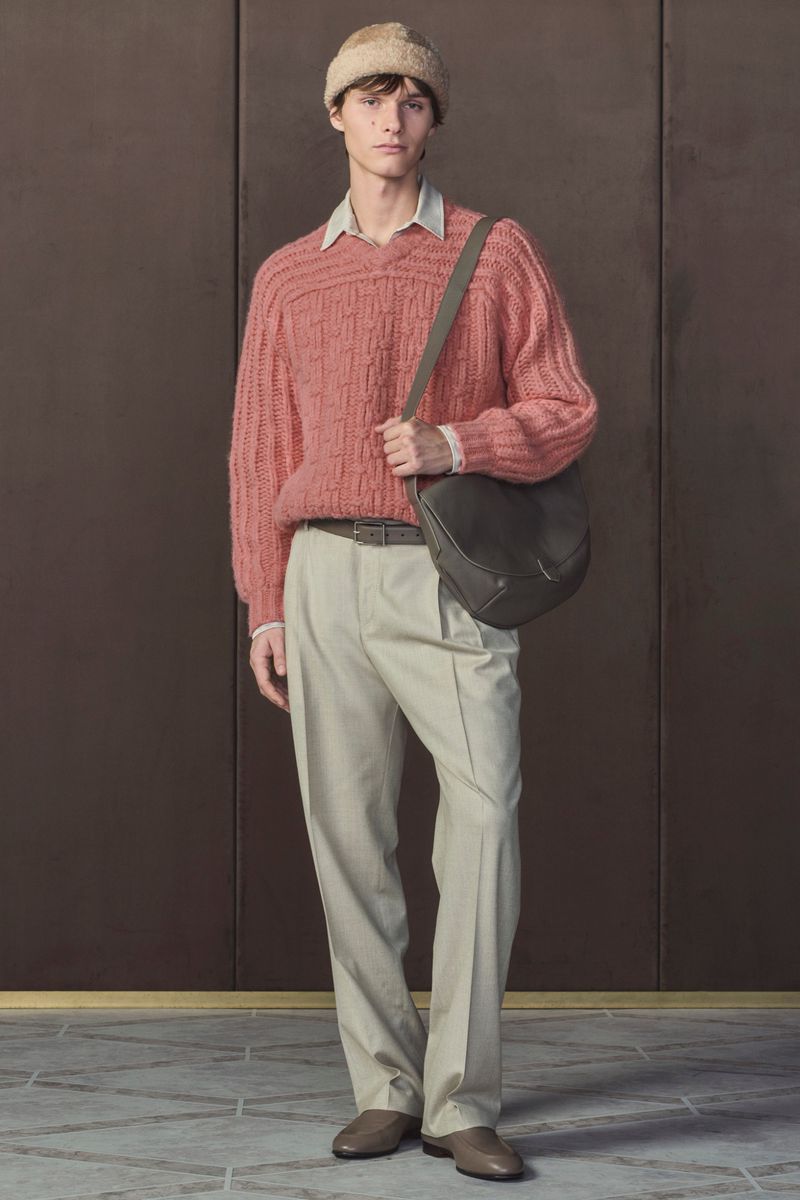 Full-length shot of a young adult man in a pink sweater, beige trousers, and a hat, accessorized with a crossbody bag against a brown background.