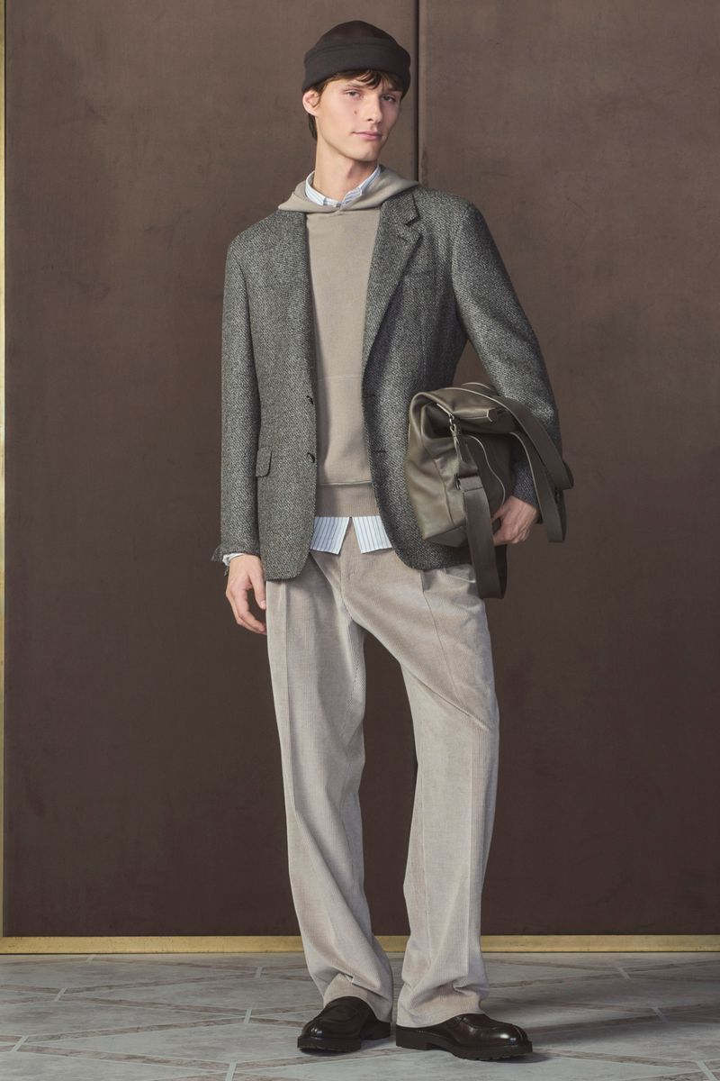 Man in a tweed jacket and corduroy trousers poses indoors holding a bag, showcasing fashion with a warm, neutral color palette.